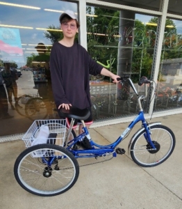 A person with disabilities stands outside a bike shop holding the handlebars of a blue adult tricycle with a white basket on the back. They are wearing sunglasses, a black long-sleeve shirt, and black shorts. The shop’s glass windows reflect bicycles and outdoor surroundings.