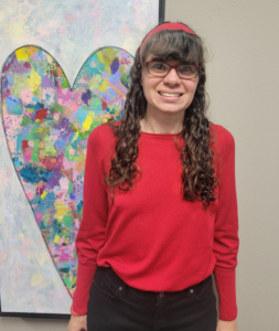 A person with long curly hair and glasses stands smiling in front of a framed painting of a colorful heart. They wear a red sweater, black pants, and a red headband against a neutral wall.