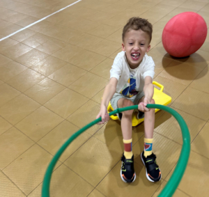 A young child sits on a yellow scooter board in a gym, gripping a green hula hoop. The child smiles with an open mouth. A red exercise ball rests on the floor behind them.